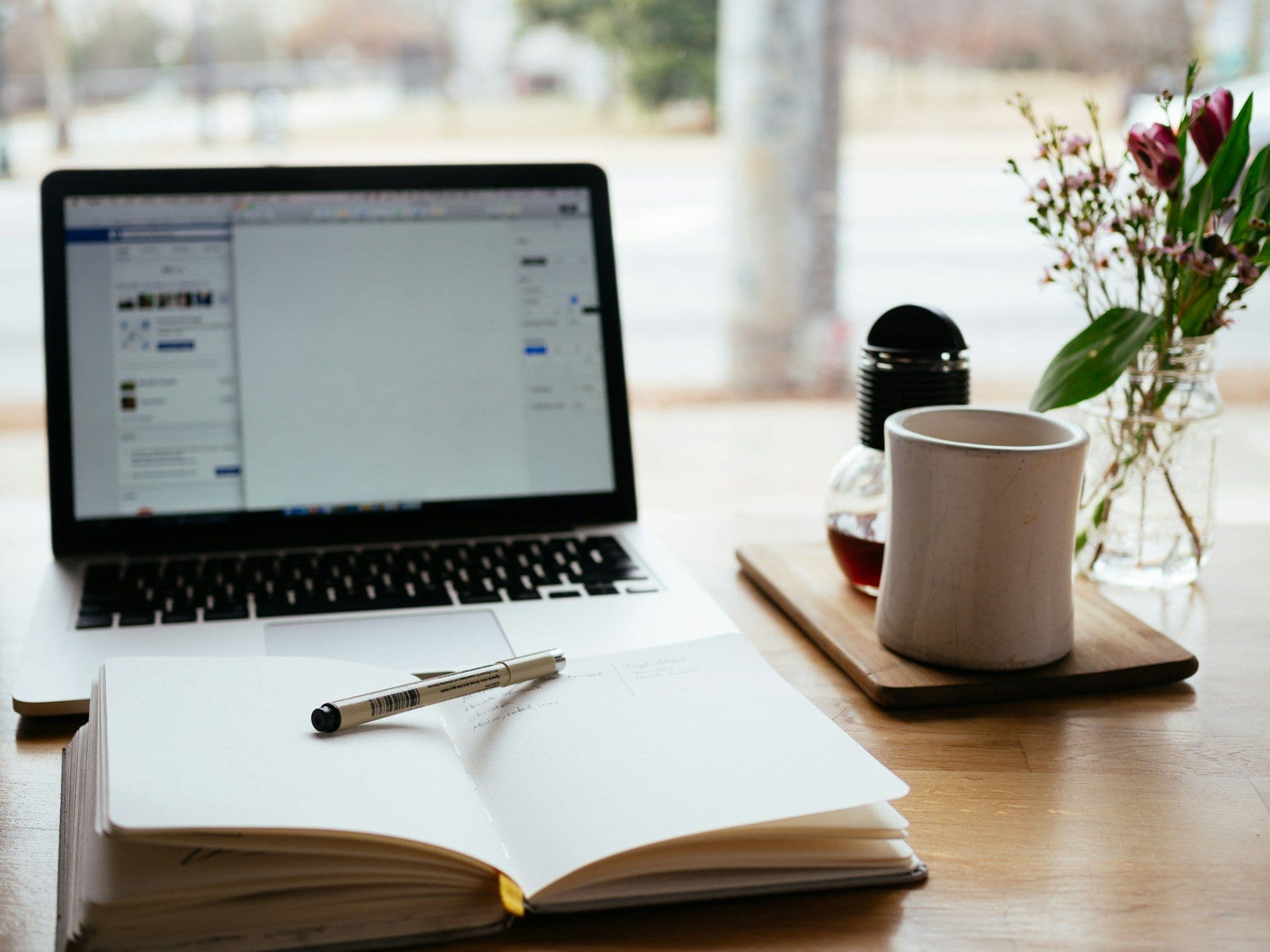 A laptop on a table by a window, with an open notebook and a pen
